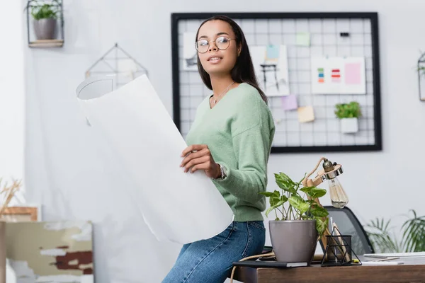 Giovane interior designer afroamericano distogliendo lo sguardo mentre tiene il progetto vicino alla scrivania a casa — Foto stock