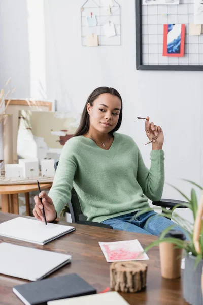 Dreamy, smiling african american interior designer holding eyeglasses and pencil while working at home, blurred foreground — Stock Photo