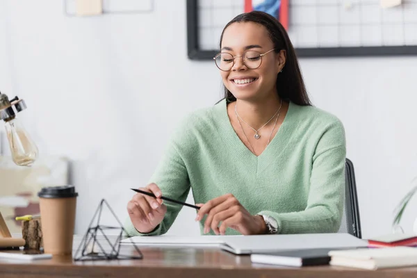 Happy african american architect in eyeglasses holding pencil while sitting at desk on blurred foreground — Stock Photo