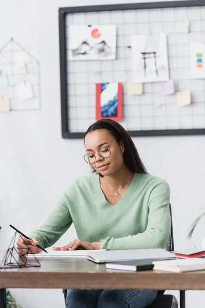 Jovem designer de interiores afro-americano desenho com lápis em estúdio em casa, foreground borrado — Fotografia de Stock