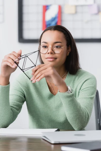 Jovem arquiteto afro-americano segurando modelo de pirâmide perto de laptop em primeiro plano borrado — Fotografia de Stock