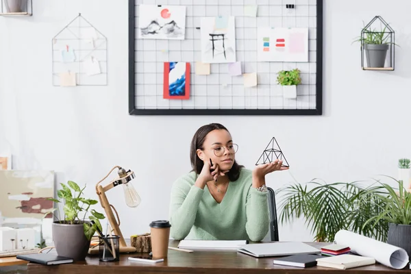 Мечтательный африканский дизайнер интерьеров держит модель пирамиды в домашней студии — стоковое фото