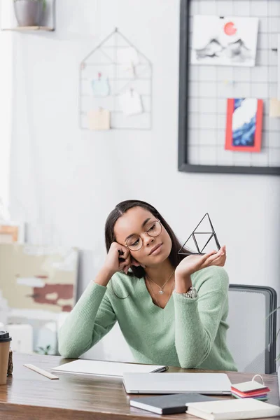 Smiling, dreamy african american interior designer hold maquette of pyramid near laptop and notebooks on blurred foreground — Stock Photo