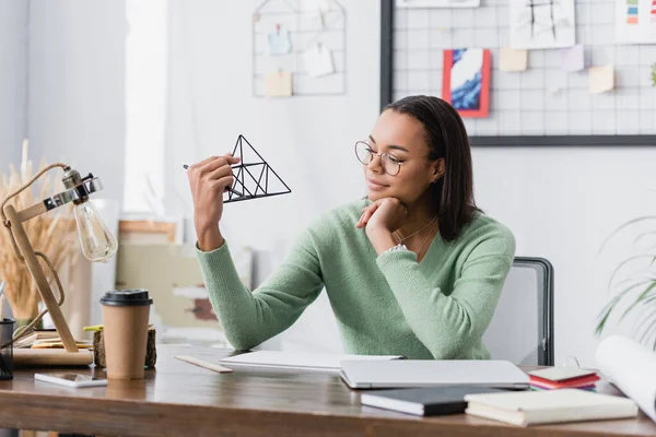 African american architect in eyeglasses holding model of pyramid near coffee to go and notebooks on blurred foreground — Stock Photo