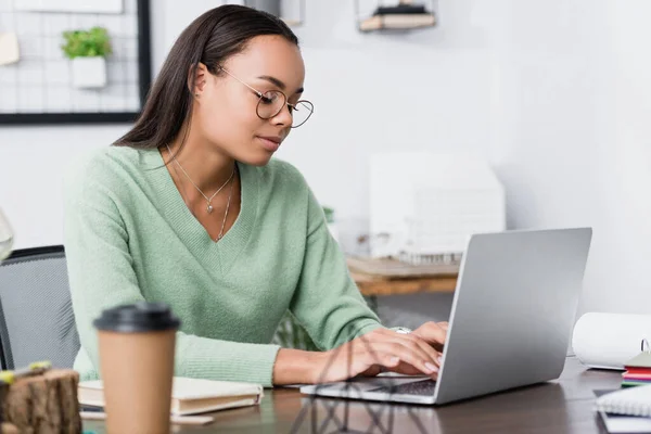 Giovane architetto afroamericano digitando sul computer portatile vicino tazza in primo piano sfocato — Foto stock