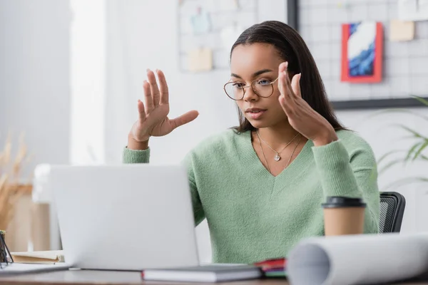 Jeune designer d'intérieur afro-américain gestuelle tout en regardant ordinateur portable sur le premier plan flou — Photo de stock