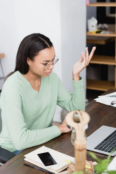 African american interior designer in eyeglasses gesturing near laptop on blurred foreground — Stock Photo
