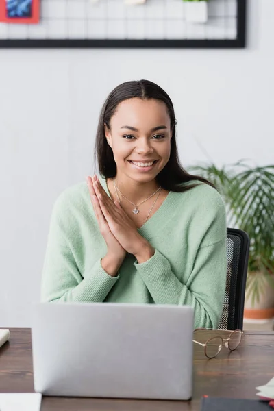 Fröhliche afrikanisch-amerikanische Innenarchitektin lächelt in die Kamera neben Laptop und Brille auf dem Schreibtisch — Stockfoto
