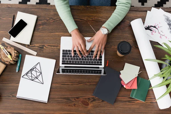 Vista recortada del arquitecto afroamericano escribiendo en el portátil cerca de teléfono inteligente, café para llevar y papelería en el escritorio - foto de stock