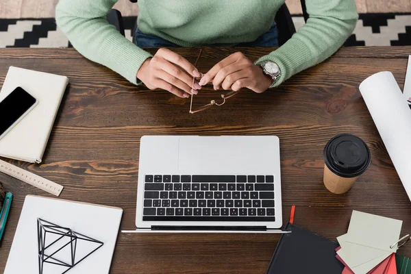 Vue de dessus du designer d'intérieur afro-américain tenant des lunettes près de l'ordinateur portable, tasse en papier et la papeterie sur le bureau — Photo de stock