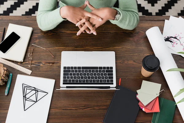 Vue partielle de l'architecte afro-américain assis avec les mains serrées près de l'ordinateur portable sur le bureau — Photo de stock