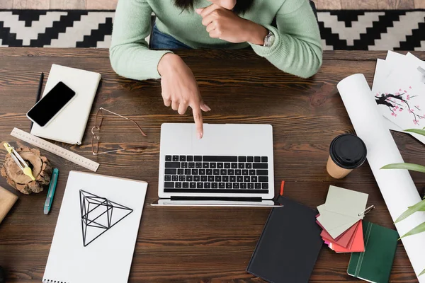 Vue du dessus du designer d'intérieur afro-américain pointant vers un ordinateur portable près du modèle pyramidal, smartphone et tasse en papier sur le bureau — Photo de stock