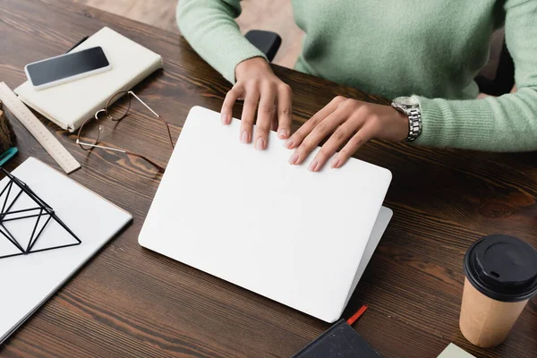 Vista parcial de la computadora portátil de apertura de diseñador de interiores afroamericano cerca del teléfono inteligente con pantalla en blanco y bebida para llevar - foto de stock