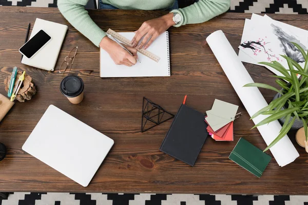 Cropped view of interior designer drawing with pencil and ruler in sketchbook near laptop, coffee to go and smartphone with blank screen — Stock Photo