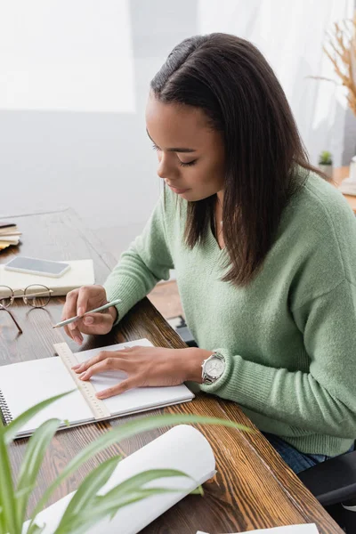 Joven diseñador afroamericano dibujo en cuaderno de bocetos con lápiz y regla en primer plano borrosa - foto de stock