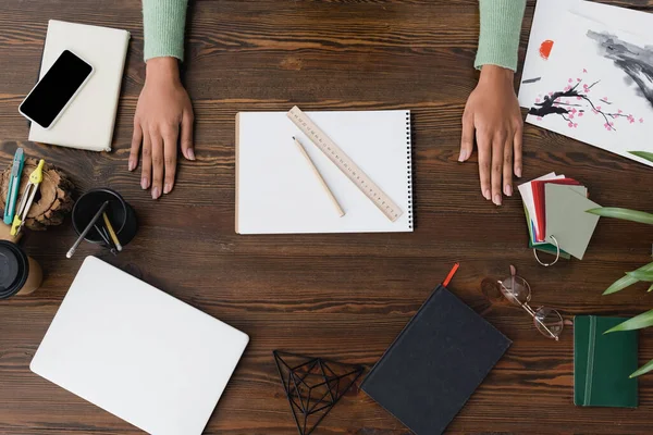 Vue partielle du designer d'intérieur afro-américain près de carnet de croquis, ordinateur portable, smartphone et papeterie sur le bureau — Photo de stock