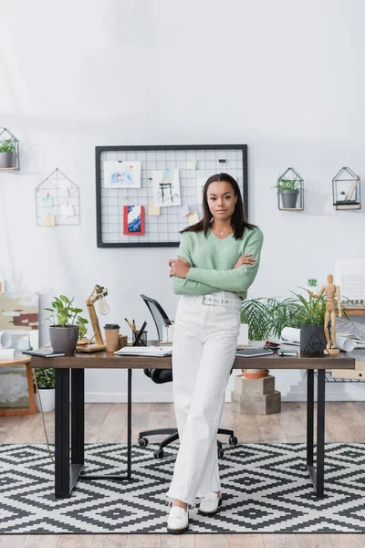 Young african american architect looking at camera while standing near desk with crossed arms — Stock Photo
