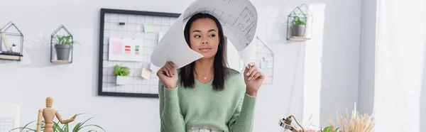 Junger afrikanisch-amerikanischer Architekt, der Kopf mit Blaupause bedeckt, während er zu Hause wegschaut, Banner — Stockfoto