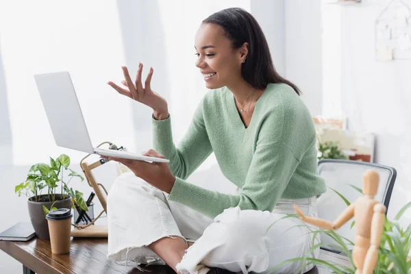 Cheerful african american freelancer sitting on desk and gesturing during video chat on laptop, blurred foreground — Stock Photo