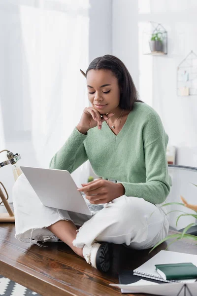 Designer d'intérieur afro-américain rêveur souriant tout en étant assis sur le bureau avec ordinateur portable — Photo de stock