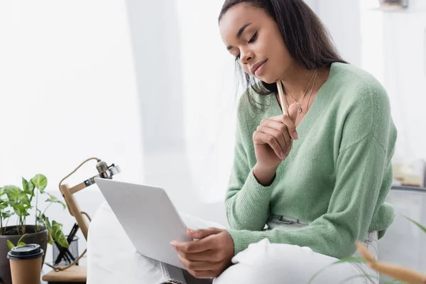 Pensativo designer de interiores afro-americano segurando lápis enquanto sentado na mesa com laptop — Fotografia de Stock