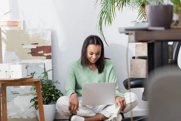 Feliz arquitecto afroamericano sonriendo mientras está sentado en el suelo con el ordenador portátil en el estudio de casa en primer plano borrosa - foto de stock