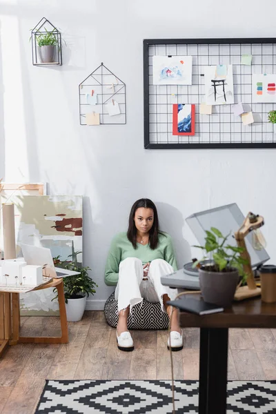 Jeune architecte afro-américain dessinant dans un carnet de croquis assis sur une chaise de sac près d'un mur avec des peintures, au premier plan flou — Photo de stock