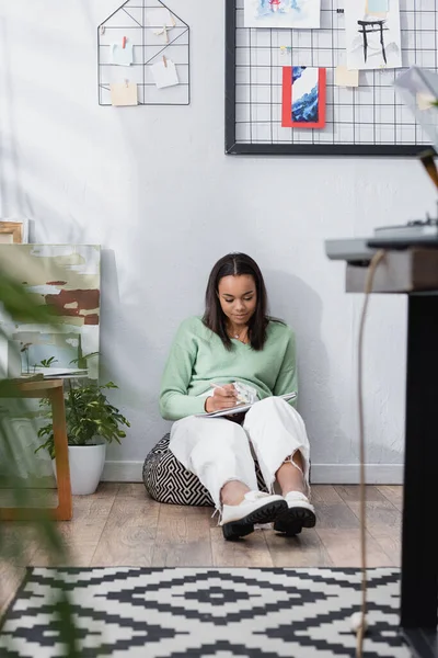 Afrikanisch-amerikanischer Innenarchitekt sitzt auf einem Stuhl in Wandnähe und zeichnet in Skizzenbuch, unscharfer Vordergrund — Stockfoto