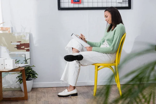 Joven arquitecto afroamericano sentado en silla en casa estudio con cuaderno de bocetos - foto de stock