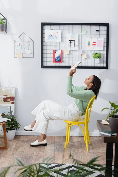 Vista laterale dell'architetto afro-americano seduto sulla sedia mentre tiene in mano sketchbook in studio a casa — Foto stock