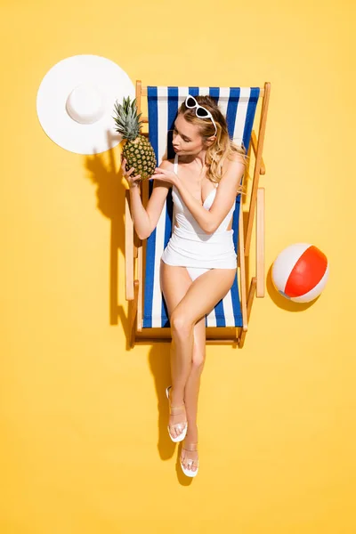 Vista dall'alto della giovane donna con ananas fresco seduto sulla sedia a sdraio vicino alla palla gonfiabile e cappello di paglia sul giallo — Foto stock