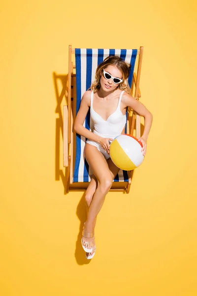 Вид сверху на молодую женщину в купальнике и солнцезащитных очках, сидящую в шезлонге с надувным мячом на желтом — стоковое фото