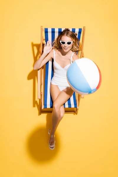 Top view of young woman in sunglasses and swimsuit waving hand while relaxing in deck chair with inflatable ball on yellow — Stock Photo