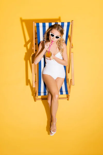 Верхний вид женщины в солнцезащитных очках и купальниках, пьющей освежающий коктейль во время отдыха в шезлонге на желтом — стоковое фото