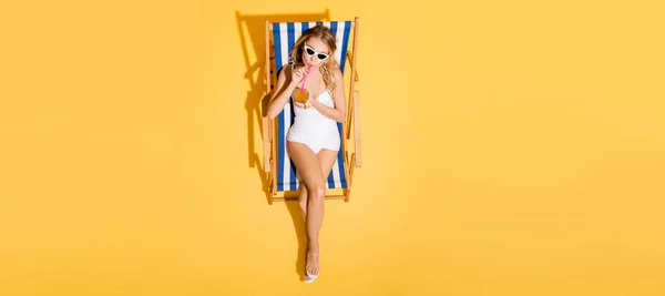 Вид зверху на жінку в купальнику і сонцезахисних окулярах, що п'ють освіжаючий коктейль в стільці на жовтому, банер — стокове фото