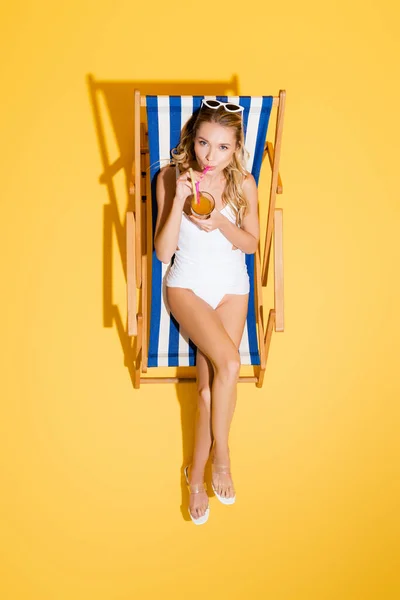 Vista superior da mulher em maiô e sandálias olhando para a câmera e beber coquetel em cadeira de praia em amarelo — Fotografia de Stock