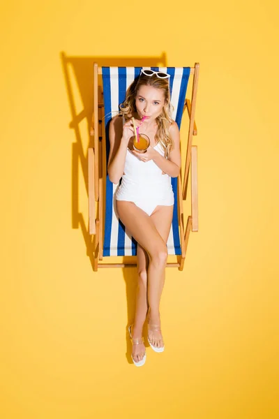 Vue du dessus de la femme en maillot de bain et sandales regardant la caméra et boire un cocktail dans la chaise longue sur jaune — Photo de stock