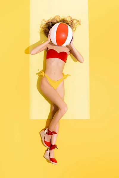 Top view of woman in swimsuit and sandals obscuring face with inflatable ball on yellow — Stock Photo