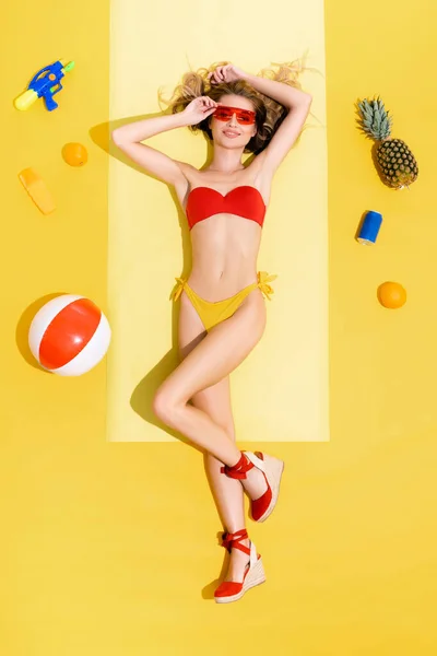 Вид зверху усміхненої жінки, що відпочиває на пляжному килимку біля фруктів, надувний м'яч, сонцезахисний крем, балончик з содовою та водяною гарматою на жовтому — стокове фото