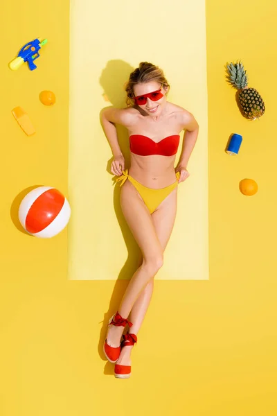 Top view of cheerful woman in swimsuit near water gun, sunscreen, inflatable ball, can of soda and fruits on yellow — Stock Photo