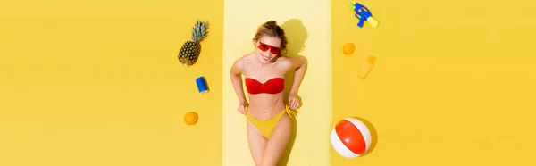Вид зверху на жінку в купальнику біля фруктів, водяний пістолет, балончик з содовою, сонцезахисний крем і надувний м'яч на жовтому, банер — стокове фото