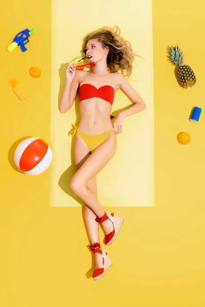 Top view of young woman holding eyeglasses while lying on beach mat near fruits, inflatable ball, water gun and sunscreen on yellow — стоковое фото