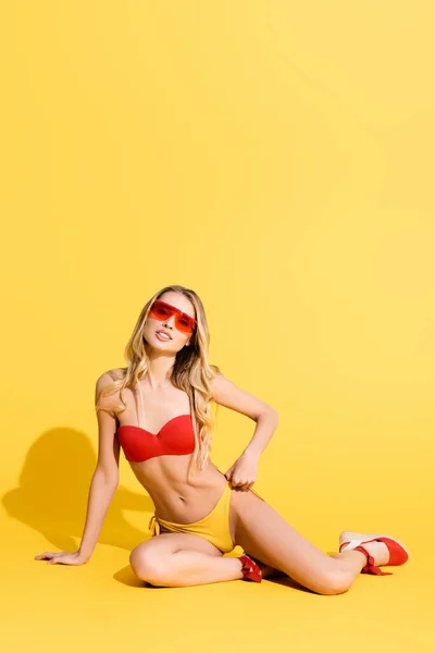 Kokettierende Frau in Sonnenbrille und Badehose blickt in die Kamera auf gelb — Stockfoto
