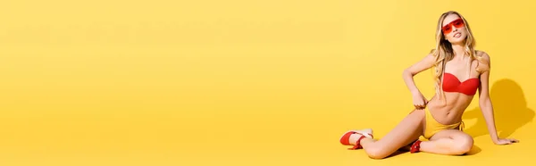 Flirty donna in costume da bagno e occhiali da sole guardando la fotocamera mentre seduto con mano sul fianco su giallo, banner — Foto stock