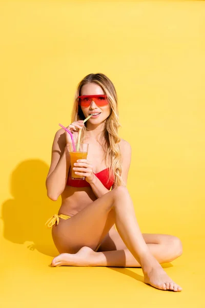Молодая босиком женщина в купальнике смотрит в камеру и пьет освежающий коктейль на желтом — стоковое фото