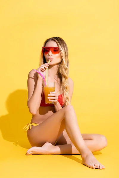 Босоногая женщина в солнцезащитных очках и купальниках пьет освежающий коктейль, глядя на желтый цвет — стоковое фото