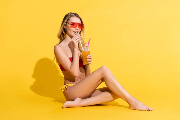 Улыбающаяся босиком женщина в купальнике смотрит в сторону и пьет свежий коктейль на желтом — стоковое фото