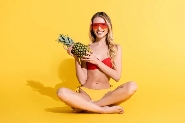 Cheerful woman in swimsuit and sunglasses holding whole pineapple while sitting with crossed legs on yellow — Stock Photo