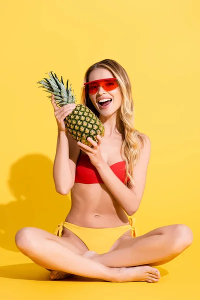 Excited woman in swimsuit and sunglasses holding pineapple while sitting with crossed legs on yellow — Stock Photo