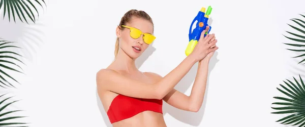 Молода жінка в жовтих сонцезахисних окулярах позує з водяним пістолетом біля пальмового листя на білому, банер — стокове фото